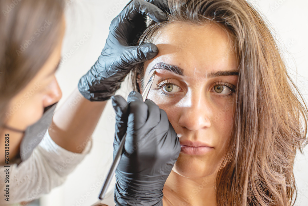 Fototapeta premium Beauty master painter paints eyebrows by coloring with henna. Woman makes a make-up in black gloves. Brow architecture