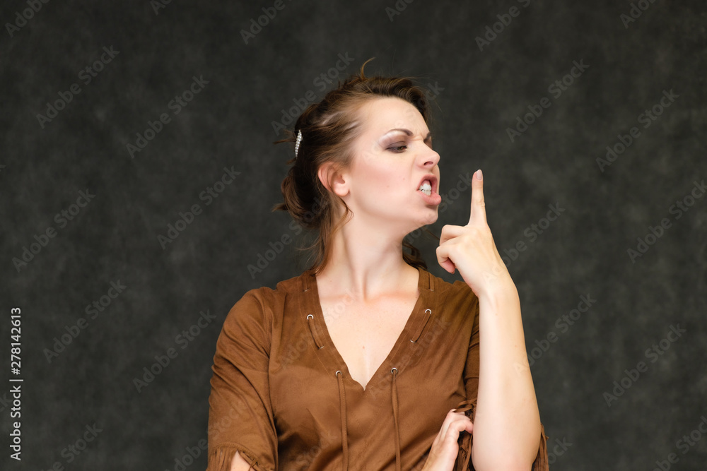 Obraz premium Portrait below breast over gray background of pretty young brunette woman in brown dress with beautiful hair. Standing in different poses, talking, showing with hands, demonstrating emotions.