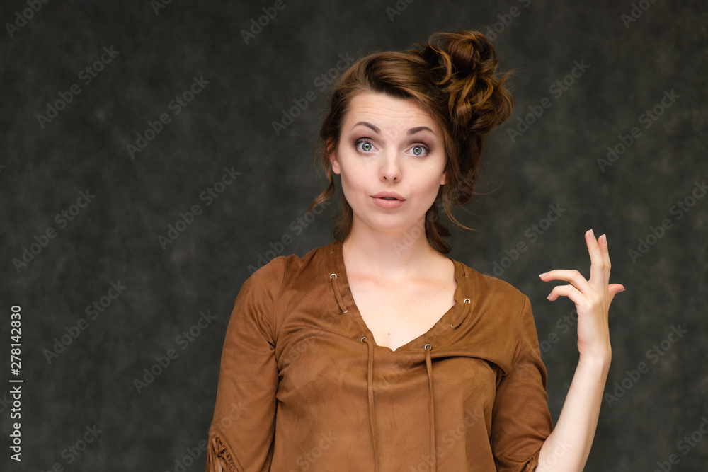 Portrait below breast over gray background of pretty young brunette woman  in brown dress with beautiful hair. Standing in different poses, talking,  showing with hands, demonstrating emotions. Stock Photo