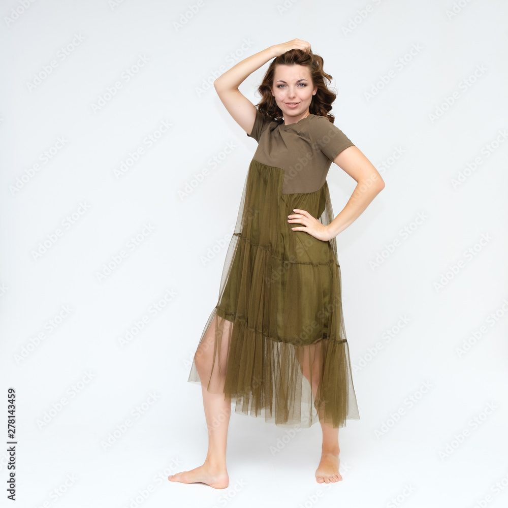 Fototapeta premium Portrait in full growth on a white background pretty young brunette woman in a green dress with beautiful hair. Standing in different poses, talking, showing hands, demonstrating emotions.