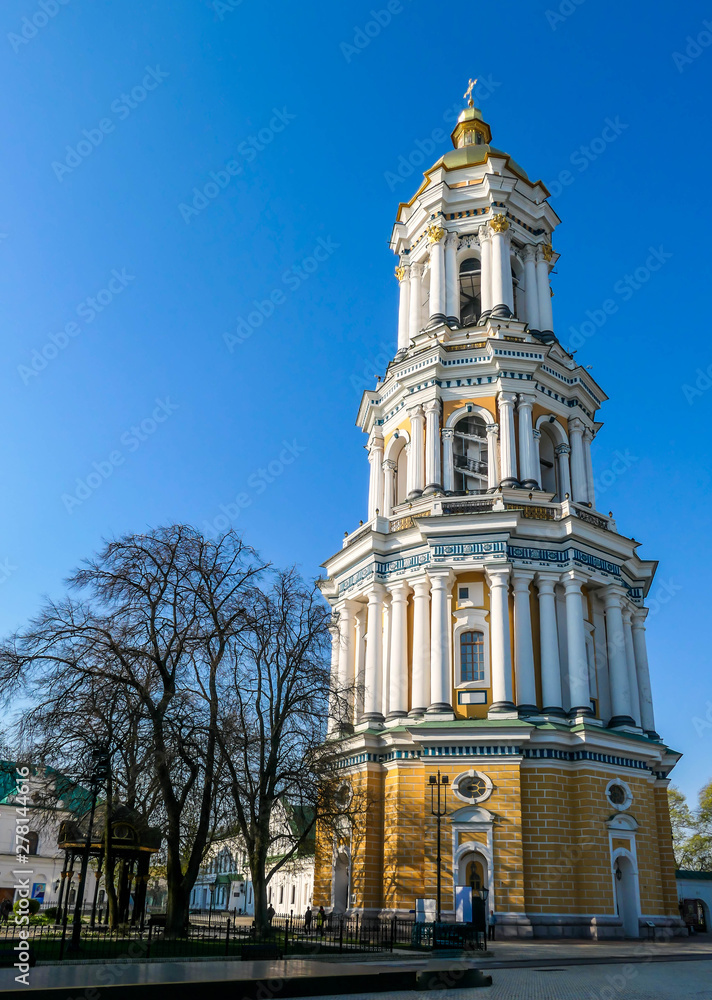 A four storied bell tower of Pechersk Lavra complex in Kiev, Ukraine. The tower is golden an white. Fe trees growing around it. Clear blue sky.