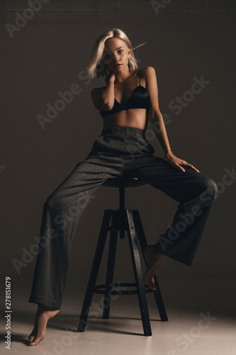 Stylish fashion trendy blonde girl with elegant hairstyle in classic trousers and black lingerie posing on chair on gray background. Beautiful hot sexy portrait of young woman model underwear in room
