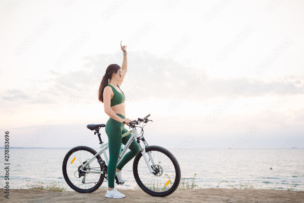 Fototapeta premium Beautiful young brunette woman resting after bicycle ride at sunrise. Model listening to music with wireless earbuds in the morning.