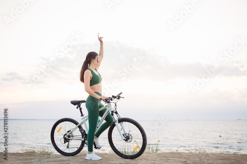 Beautiful young brunette woman resting after bicycle ride at sunrise. Model listening to music with wireless earbuds in the morning.