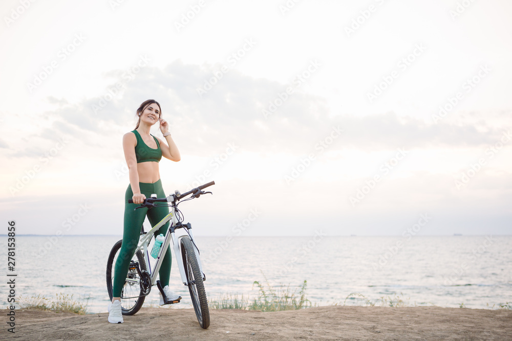 Fototapeta premium Beautiful young brunette woman resting after bicycle ride at sunrise. Model listening to music with wireless earbuds in the morning.