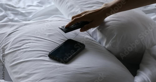Woman's hand using TV remote control and smartphone on bed in the morning