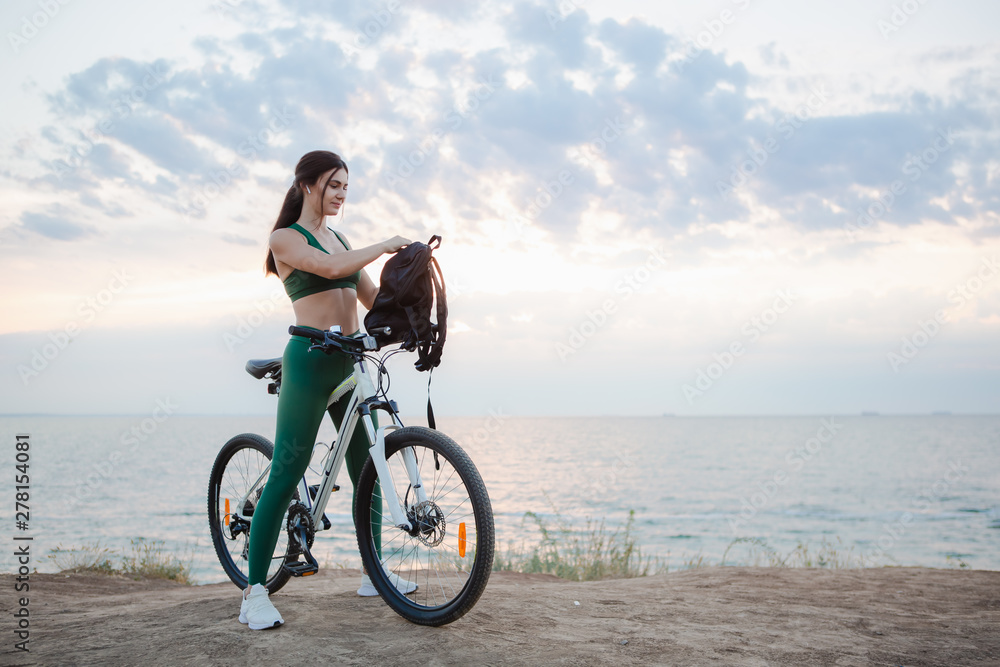 Young brunette woman with a backpack and bicycle rest at the sea shore in the morning after cycling. Young brunette woman with a backpack and bicycle rest at the sea shore in the morning after cycling.