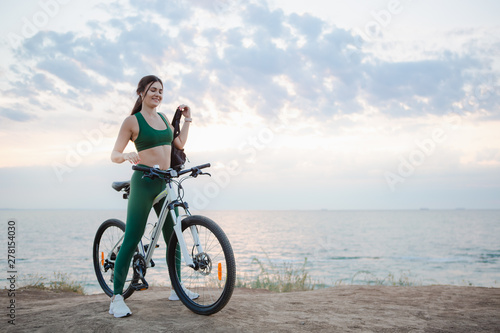 Young brunette woman with a backpack and bicycle rest at the sea shore in the morning after cycling.
