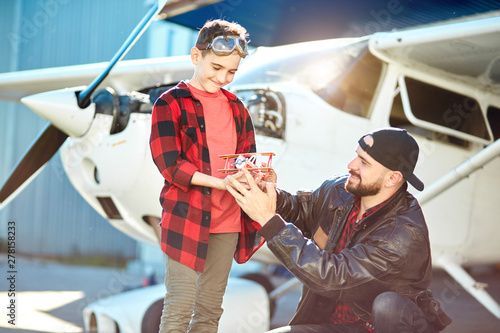 view of son and father holding toy plane that they made together, smiling happily and enjoying their aeromodelling hobby, outdoor shot. Aircraft and family concept. photo