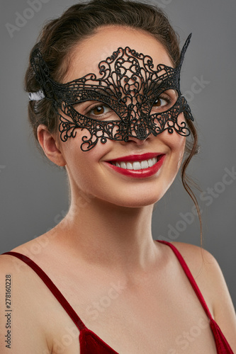Half-turn shot of smiling lady with dark hair, wearing wine red crop top. The woman is tilting her head and looking at camera, wearing black carnival mask with perforation, tied with satin ribbon.