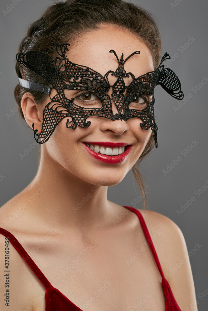 Obraz premium Half-turn shot of woman with tied back dark hair, wearing wine red crop top. The smiling lady is tilting her head, wearing black masquerade mask with fancy perforation. Vintage carnival accessory.