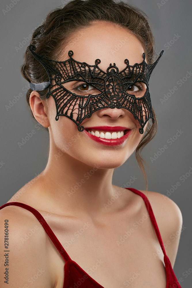 Half-turn shot of girl with dark hair, wearing wine red crop top. The  smiling lady is looking at camera, wearing black butterfly-shaped  masquerade mask with perforation. Vintage carnival accessory. Stock Photo