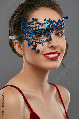 Half-turn shot of woman with dark hair, wearing wine red crop top. The smiling girl is looking at camera, wearing blue asymmetric carnival mask with perforation. Vintage women's carnival accessory. photo