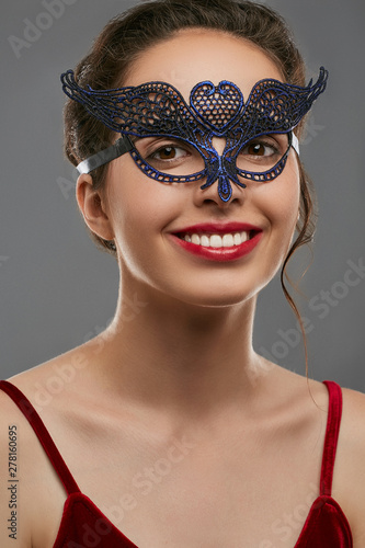 Half-turn portrait of smiling woman with dark hair, wearing wine red crop top. The young girl is looking at camera, wearing blue carnival mask with fancy perforation in view of heart in forehead. photo