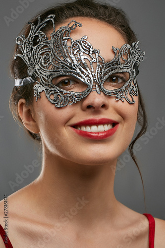 Half-turn shot of smiling lady with tied back dark hair, wearing wine red crop top. The woman is raising her head, wearing asymmetric silver carnival mask with perforation, tied with satin ribbon.