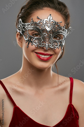 Front portrait of smiling woman with tied back dark hair, wearing wine red crop top. The young girl is tilting head, wearing silver opalesce carnival mask with fancy perforation and jutting edges.