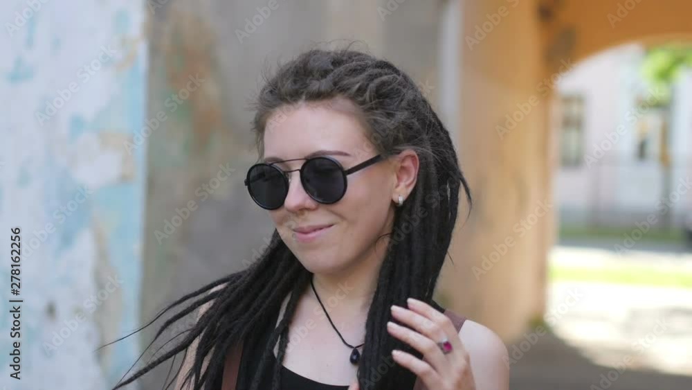 Portrait of an attractive girl with dreadlocks in sunglasses standing and smiling on a city street Portrait of an attractive girl with dreadlocks in sunglasses standing and smiling on a city street