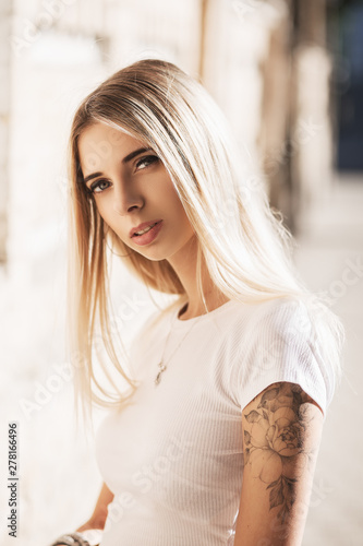  Young blond woman with flower tattoo wearing white t-shirt. She is looking at camera. Fashion portrait. Close-up.
