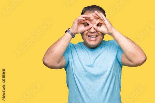 Middle age arab man wearing blue t-shirt over isolated background doing ok gesture like binoculars sticking tongue out, eyes looking through fingers. Crazy expression.