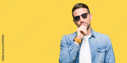 Handsome man wearing fashion sunglasses with hand on chin thinking about question, pensive expression. Smiling with thoughtful face. Doubt concept.