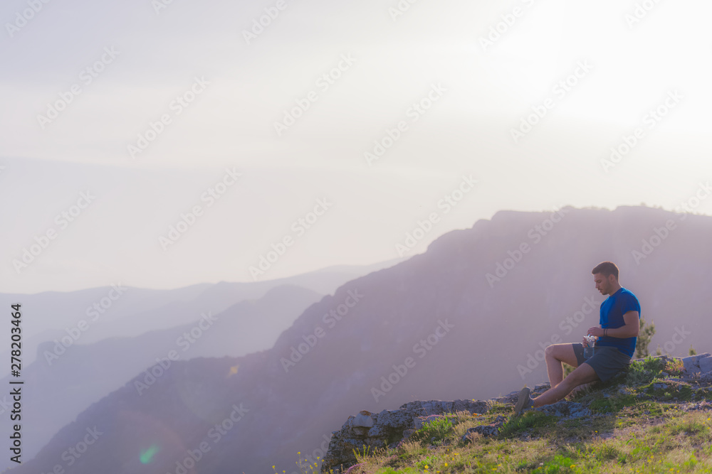 Handsome athlete sitting at a rocky peak while looking at the breathtaking mountain line and a beautiful lake while wearing a blue shirt and grey shorts.