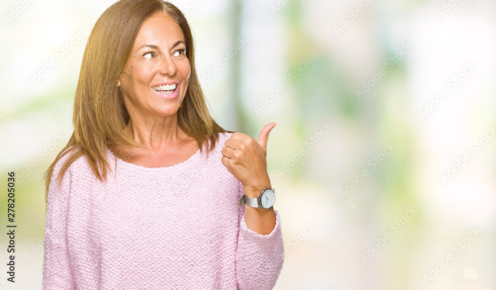 Beautiful middle age adult woman wearing winter sweater over isolated background smiling with happy face looking and pointing to the side with thumb up. Beautiful middle age adult woman wearing winter sweater over isolated background smiling with happy face looking and pointing to the side with thumb up.