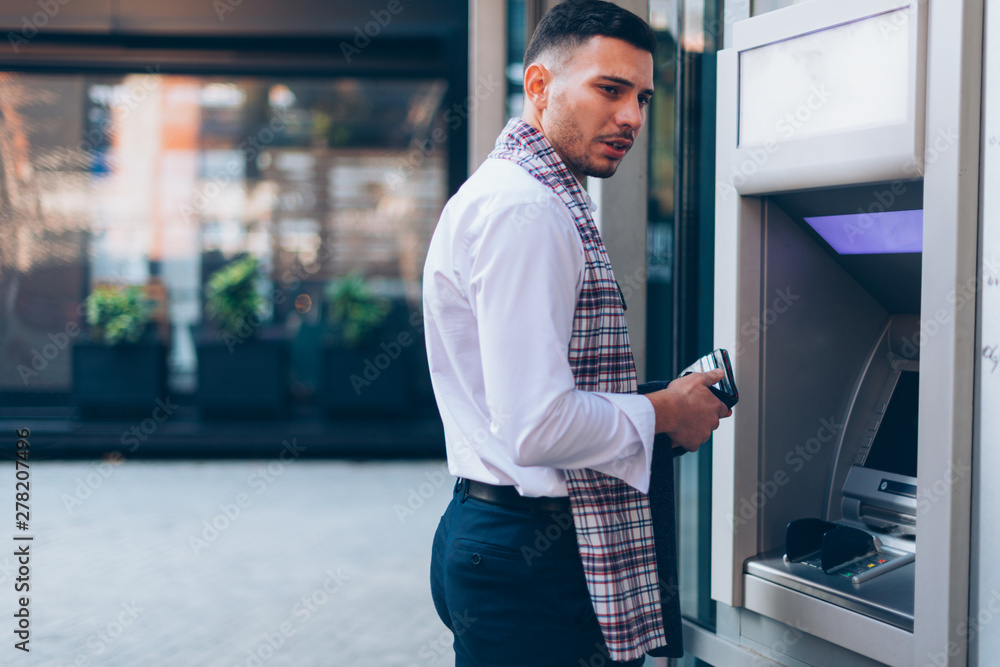 Stylish businessman in white shirt is holding his wallet and standing next to the automated teller machine. Stylish businessman in white shirt is holding his wallet and standing next to the automated teller machine.
