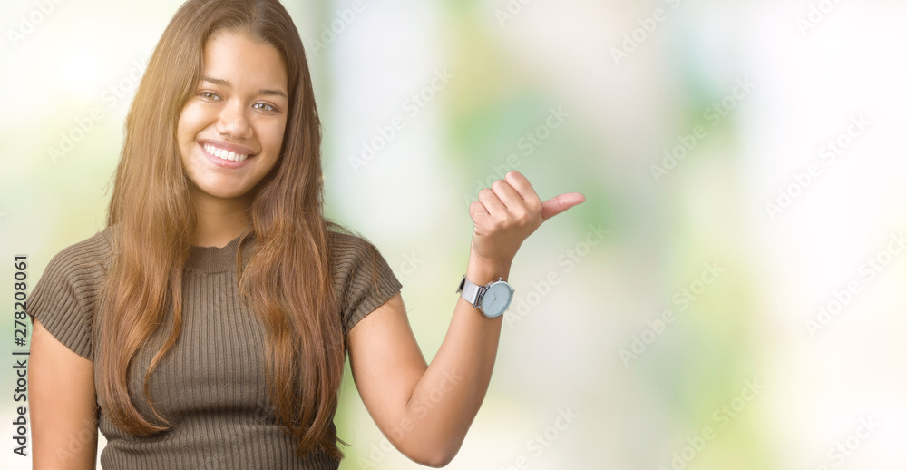 Young beautiful brunette woman over isolated background smiling with happy face looking and pointing to the side with thumb up. Young beautiful brunette woman over isolated background smiling with happy face looking and pointing to the side with thumb up.