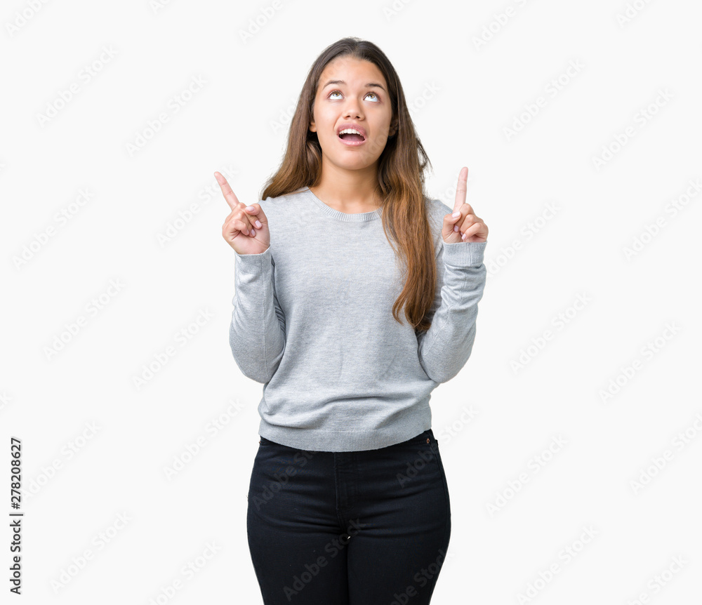 Young beautiful brunette woman wearing sweater over isolated background amazed and surprised looking up and pointing with fingers and raised arms. Young beautiful brunette woman wearing sweater over isolated background amazed and surprised looking up and pointing with fingers and raised arms.
