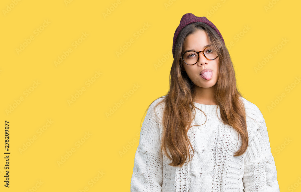 Young beautiful brunette hipster woman wearing glasses and winter hat over isolated background sticking tongue out happy with funny expression. Emotion concept.