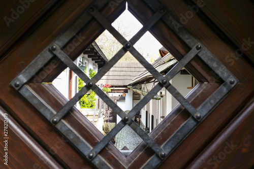 View of cozy old courtyard through rhombus window with cells in gate. Medieval narrow cobbled street and small ancient houses in Novy Svet, Hradcany district. Young greenery on white walls. Nobody