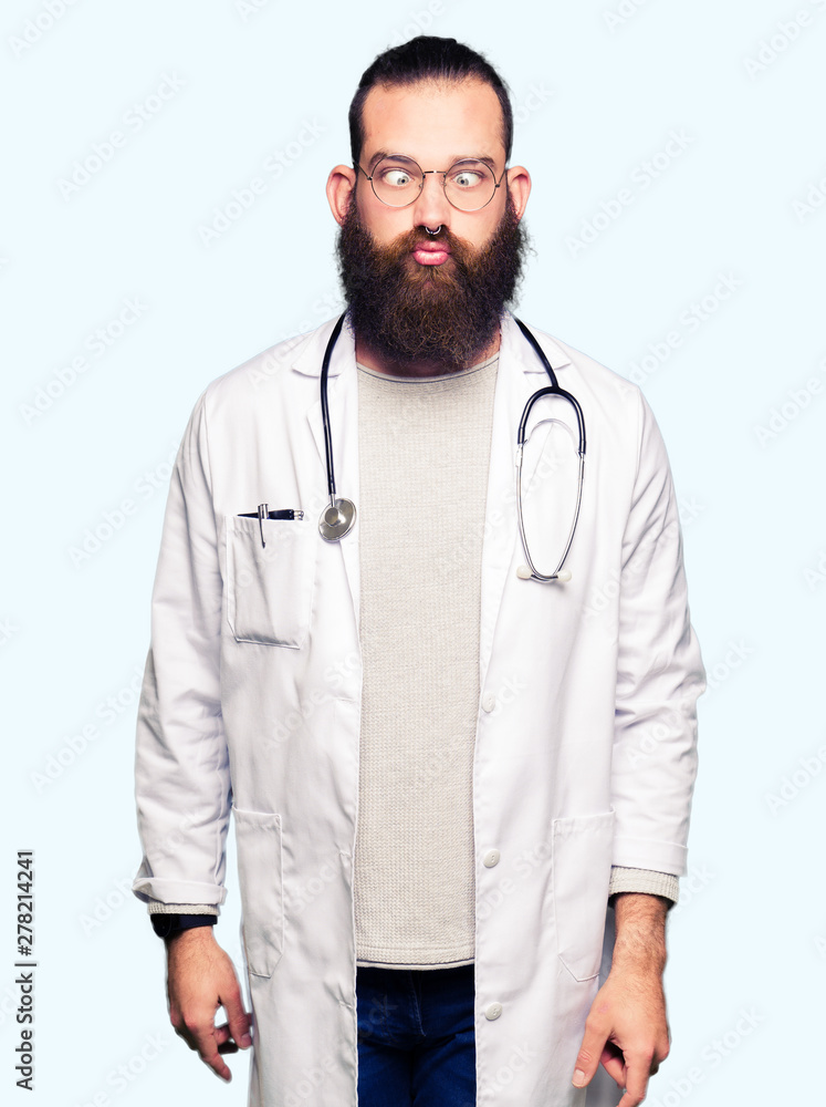Young blond doctor man with beard wearing medical coat making fish face with lips, crazy and comical gesture. Funny expression. Young blond doctor man with beard wearing medical coat making fish face with lips, crazy and comical gesture. Funny expression.