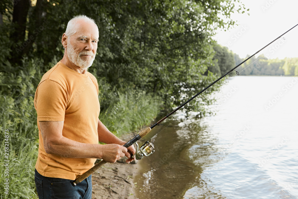 Age, activity and leisure concept. Side view of retired senior bearded male feeling relaxed and happy while fishing on river bank with fisher rod cast in water, waiting for fish to be hooked Age, activity and leisure concept. Side view of retired senior bearded male feeling relaxed and happy while fishing on river bank with fisher rod cast in water, waiting for fish to be hooked