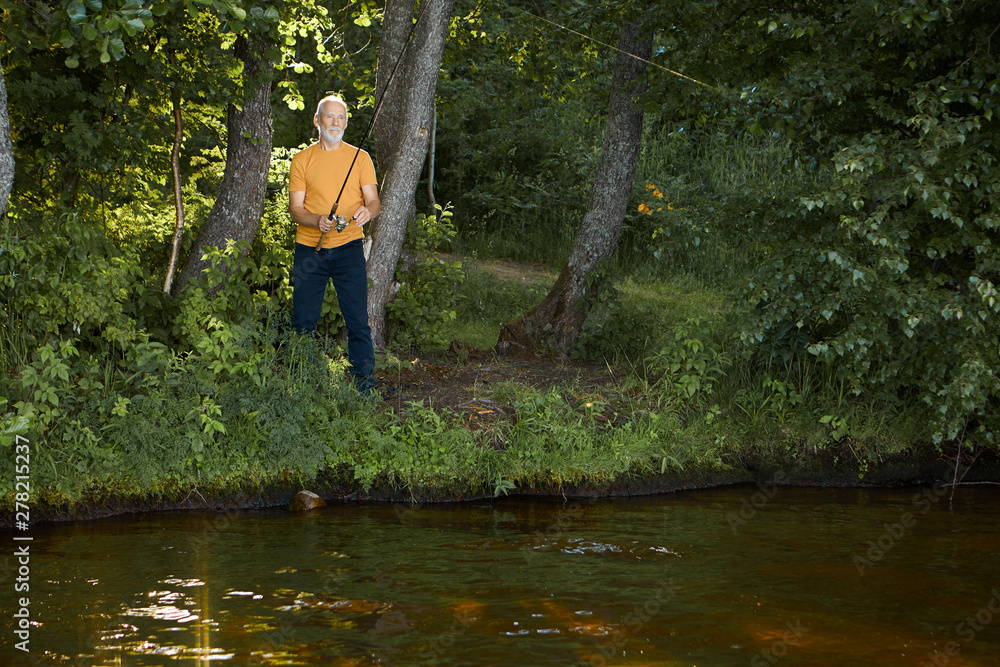 Horizontal outdoor shot of serious healthy elderly man pensioner standing in bushes on river bank, holding angler or fishing rod cast in water. Angling, fishery and recreation. View from opposite side