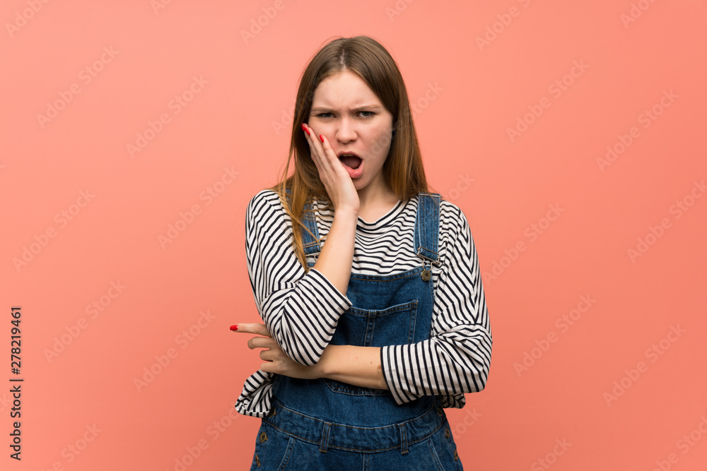 Young woman with overalls over pink wall surprised and shocked while looking right Young woman with overalls over pink wall surprised and shocked while looking right