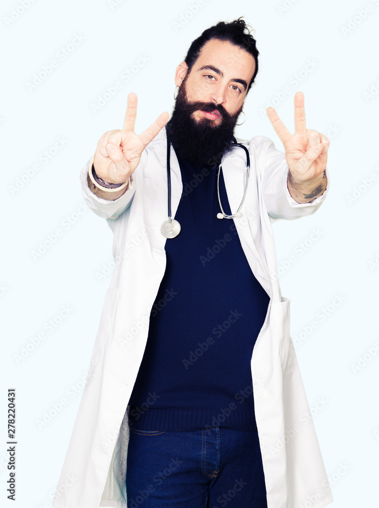 Doctor with long hair wearing medical coat and stethoscope smiling looking to the camera showing fingers doing victory sign. Number two. Doctor with long hair wearing medical coat and stethoscope smiling looking to the camera showing fingers doing victory sign. Number two.