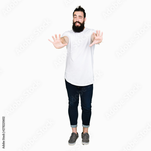 Young hipster man with long hair and beard wearing casual white t-shirt afraid and terrified with fear expression stop gesture with hands, shouting in shock. Panic concept. © Krakenimages.com