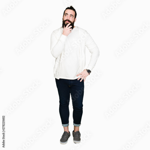 Young man with long hair and beard wearing sporty sweatshirt with hand on chin thinking about question, pensive expression. Smiling with thoughtful face. Doubt concept.