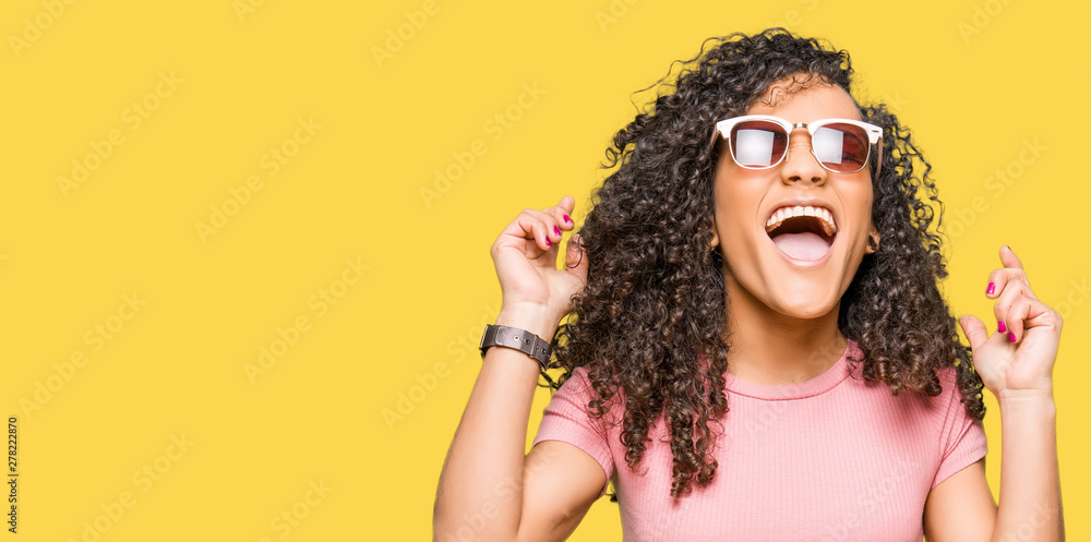 Young beautiful woman with curly hair wearing pink sunglasses celebrating mad and crazy for success with arms raised and closed eyes screaming excited. Winner concept Young beautiful woman with curly hair wearing pink sunglasses celebrating mad and crazy for success with arms raised and closed eyes screaming excited. Winner concept