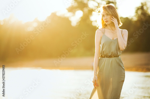 Young beautiful slim woman with short light hair wearing green summer dress walking at sea coast at sunset.