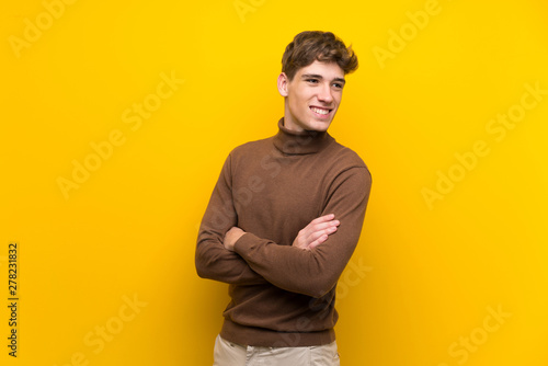 Handsome young man over isolated yellow background with arms crossed and happy