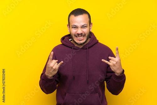 Colombian man with sweatshirt over yellow wall making rock gesture
