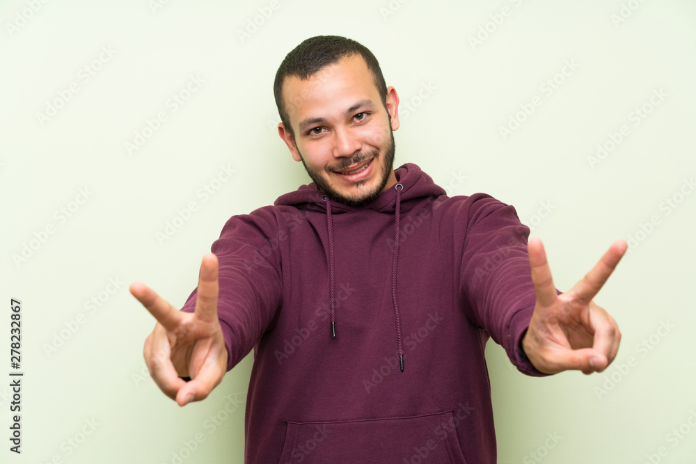 Colombian man with sweatshirt over green wall smiling and showing victory sign Colombian man with sweatshirt over green wall smiling and showing victory sign