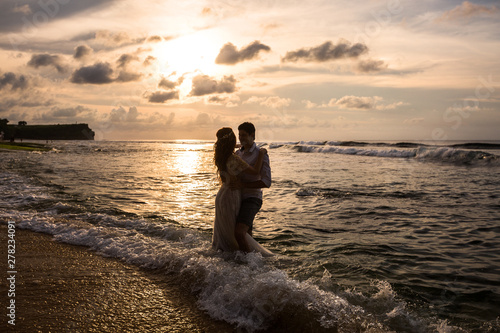 Young couple embracing standing in the sea water on the beach at sunset. Newlywed happy young couple embracing enjoying ocean sunset