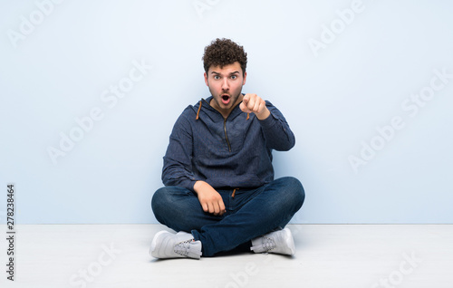 Young man sitting on the floor surprised and pointing front © luismolinero