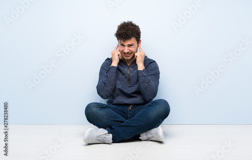 Young man sitting on the floor frustrated and covering ears © luismolinero