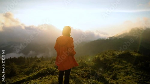 hipster millennial young woman in orange jacke on top of mountain looking at sunset view