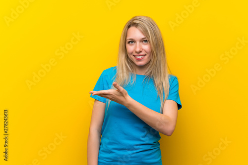 Young blonde woman over isolated yellow background presenting an idea while looking smiling towards