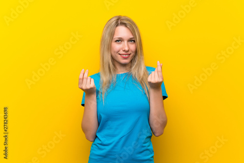 Young blonde woman over isolated yellow background making money gesture