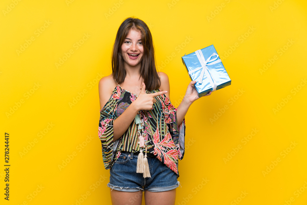Caucasian girl in colorful dress over isolated yellow background holding gift box Caucasian girl in colorful dress over isolated yellow background holding gift box